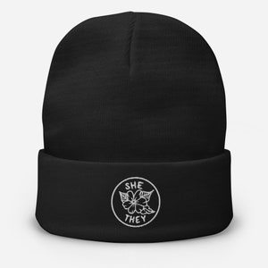 May include: Black beanie with a white embroidered circle containing the words "SHE THEY" and a stylised flower.