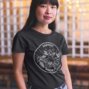May include: A black short-sleeved t-shirt featuring a white circular graphic print. The design depicts two hands reaching toward each other surrounded by flowers and stars, with the text SAPPHIC SOCIETY arched along the top and bottom edges.