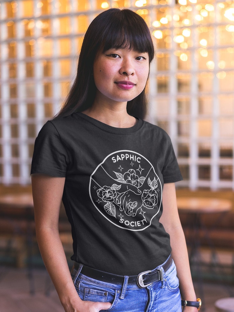 May include: A black short-sleeved t-shirt featuring a white circular graphic print. The design depicts two hands reaching toward each other surrounded by flowers and stars, with the text SAPPHIC SOCIETY arched along the top and bottom edges.