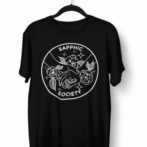 May include: A black short-sleeve t-shirt featuring a white circular graphic on the chest. The design depicts two hands reaching toward each other surrounded by flowers and small star icons, with the text SAPPHIC SOCIETY printed in white.