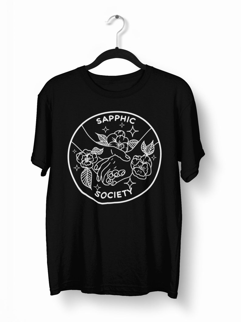 May include: A black short-sleeve t-shirt featuring a white circular graphic on the chest. The design depicts two hands reaching toward each other surrounded by flowers and small star icons, with the text SAPPHIC SOCIETY printed in white.