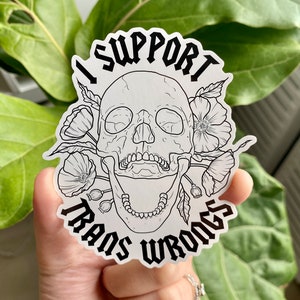 以下が含まれることがあります： 骸骨と花が描かれた白黒のステッカー。"I support trans wrongs"というテキストがゴシック体で書かれています。