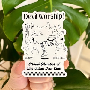 May include: Black and white sticker with a cartoon heart-shaped devil holding a pitchfork. The text "Devil Worship!" is at the top of the sticker. The text "Be Gay" and "Raise Hell" are at the bottom of the sticker. The text "Proud Member of The Satan Fan Club" is in the middle of the sticker.