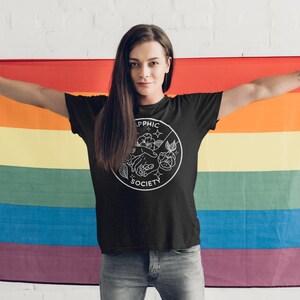 May include: A black short-sleeved t-shirt featuring a white circular graphic on the chest. The design includes the text "SAPPHIC SOCIETY" with an illustration of two hands touching, surrounded by floral elements and small star shapes.