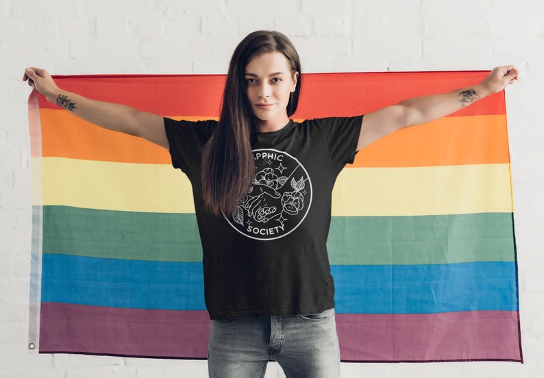 May include: A black short-sleeved t-shirt featuring a white circular graphic on the chest. The design includes the text "SAPPHIC SOCIETY" with an illustration of two hands touching, surrounded by floral elements and small star shapes.