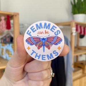 Puede incluir: Una pegatina redonda blanca con texto azul que dice "Femmes can be Them" y una ilustración de mariposa colorida.