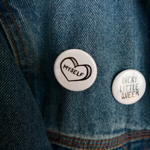 May include: Two white button badges on a blue denim jacket. The left badge has a black heart with the word "myself" inside. The right badge has the words "lucky little queer" in black text.
