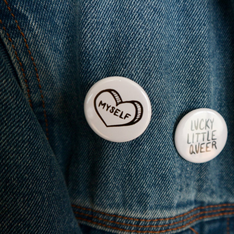 May include: Two white button badges on a blue denim jacket. The left badge has a black heart with the word "myself" inside. The right badge has the words "lucky little queer" in black text.