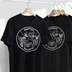 May include: A black t-shirt featuring a white circular graphic with the text "SAPPHIC SOCIETY" surrounding an illustration of two hands reaching toward each other, accented by floral elements and small four-pointed star shapes.