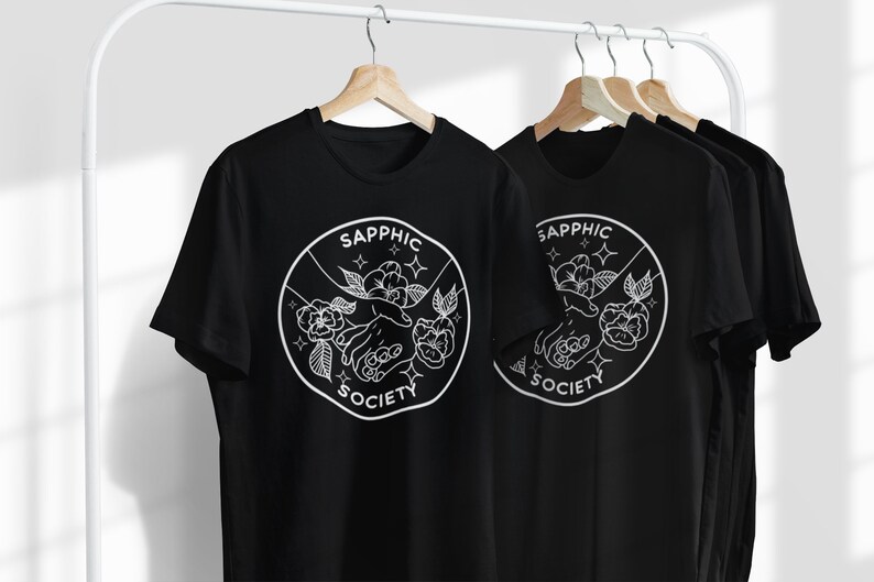 May include: A black t-shirt featuring a white circular graphic with the text "SAPPHIC SOCIETY" surrounding an illustration of two hands reaching toward each other, accented by floral elements and small four-pointed star shapes.