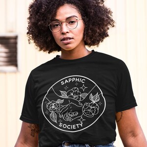 May include: A black short-sleeve t-shirt featuring a white circular graphic print. The design depicts two hands reaching toward each other surrounded by flowers and small star icons, with the text SAPPHIC SOCIETY arched along the border.