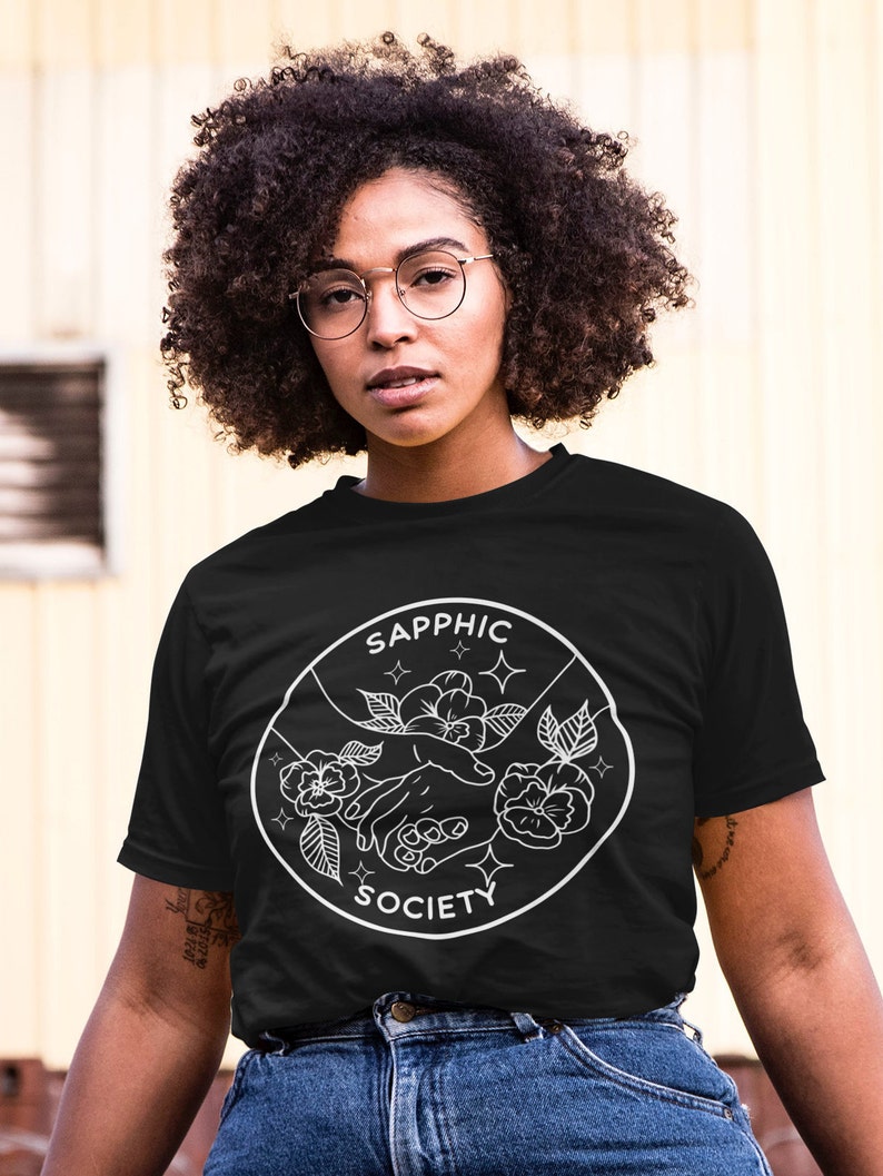 May include: A black short-sleeve t-shirt featuring a white circular graphic print. The design depicts two hands reaching toward each other surrounded by flowers and small star icons, with the text SAPPHIC SOCIETY arched along the border.