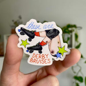 Könnte beinhalten: Ein Aufkleber mit einer Cartoon-Illustration von Beinen mit Rollschuhen und Prellungen. Der Text "these are" steht in blauer Schrift über den Beinen, und "DERBY BRUISES" steht in roten Blockbuchstaben darunter. Der Aufkleber hat einen weißen Hintergrund mit gelben und blauen Sternakzenten.