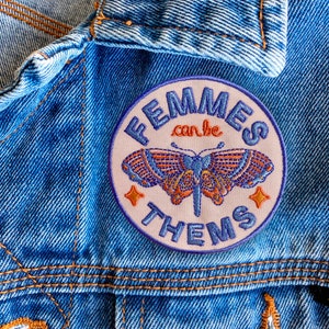 Pode incluir: Um remendo bordado rosa e azul com uma borboleta e o texto "FEMMES can be THEMS".