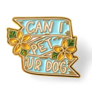 May include: A gold-colored enamel pin with a light blue banner design. The pin features the text "CAN I PET UR DOG" in white lettering, with yellow flowers and green leaves. The pin is a decorative accessory.