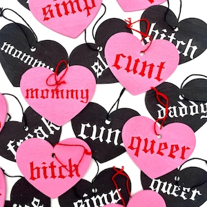 Peut inclure: Ornements en forme de cœur roses et noirs, avec des mots en police gothique. Les cœurs roses ont un texte rouge, les noirs un texte blanc. Chaque ornement est percé d'un trou avec une ficelle rouge.