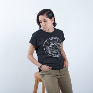 May include: A black short-sleeved t-shirt featuring a white circular graphic on the chest. The design depicts two hands holding a flower, surrounded by small stars and the text "SAPPHIC SOCIETY" written in a clean, sans-serif font.