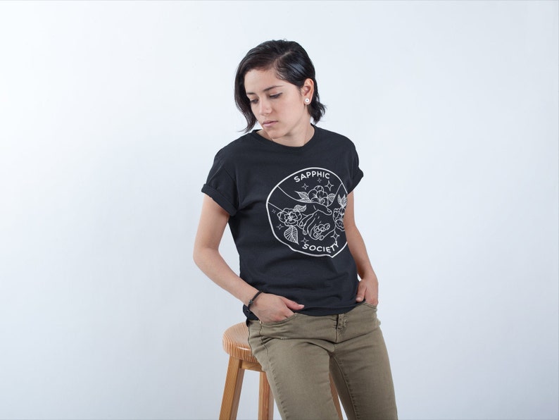 May include: A black short-sleeved t-shirt featuring a white circular graphic on the chest. The design depicts two hands holding a flower, surrounded by small stars and the text "SAPPHIC SOCIETY" written in a clean, sans-serif font.