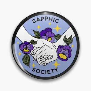 May include: A circular badge with a black edge, displaying the text "Sapphic Society". The design includes two white hands clasped, purple pansies, and yellow stars on a light blue background.