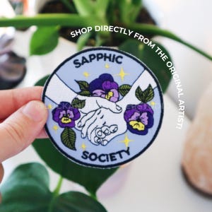 May include: A blue and white embroidered patch with the words "Sapphic Society" and two hands holding purple flowers.