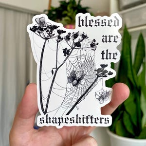 May include: A white sticker featuring a black illustration of a spiderweb with flowers and moths. The text "blessed are the shapeshifters" is printed in a gothic font. The sticker is held in a hand.