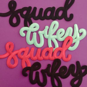 Puede incluir: Cuatro parches de tela bordados con letras cursivas. Dos parches muestran la palabra squad en negro y rosa coral, mientras que dos parches muestran la palabra wifey en verde menta y negro, todo en un estilo de escritura fluido y continuo.