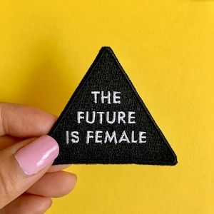 Może przedstawiać: Czarna, trójkątna naszywka z białym napisem "THE FUTURE IS FEMALE". Naszywka jest prezentowana na tle jaskrawożółtego tła.