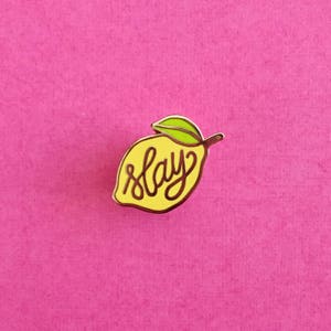 Peut inclure: Une broche en émail en forme de citron jaune avec une feuille verte sur le dessus. Le mot slay est écrit en lettres cursives marron foncé au centre du citron. La broche présente un contour en métal doré et une finition lisse et brillante.