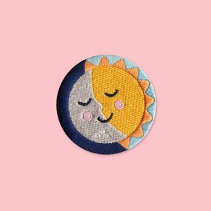 May include: Embroidered patch depicting a smiling sun and moon with closed eyes. The sun is yellow with orange rays, and the moon is silver. The patch has a blue border and is set against a pink background.