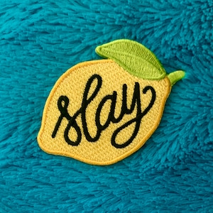 Könnte beinhalten: Ein gelber, limonenförmiger Patch mit einem grünen Blatt und dem Wort "slay" in schwarzer Kursivschrift.