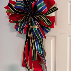 May include: A large decorative bow made from multiple ribbons in solid red, navy blue, and green, accented with metallic gold and silver stripes. The bow features numerous loops and long, flowing tails that hang downward in a layered arrangement.