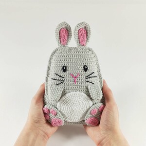 May include: A handmade gray crochet bunny backpack with pink accents on the ears and paws. The bunny has embroidered black whiskers and eyes, and a pink nose. The backpack is held by two hands.
