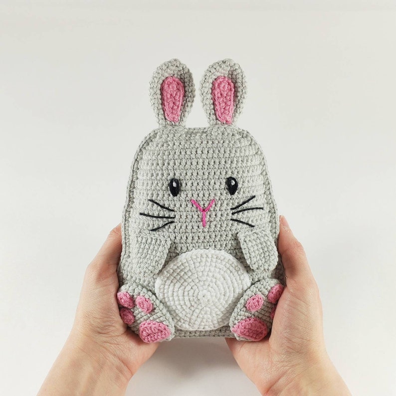 May include: A handmade gray crochet bunny backpack with pink accents on the ears and paws. The bunny has embroidered black whiskers and eyes, and a pink nose. The backpack is held by two hands.