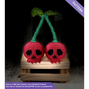 May include: Two crocheted cherries with skull faces, red bodies, green stems, and leaves. The image includes the text "PATTERN" and "This is a PDF file Tutorial, not a finished Product. Ceci est un Tutoriel au format PDF et non un produit fini."