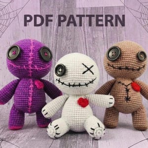 Puede incluir: Tres muñecos de ganchillo hechos a mano con ojos de botón y sonrisas cosidas. Los muñecos son de color morado, blanco y marrón, cada uno con un acento en forma de corazón. La imagen incluye el texto "PDF PATTERN" y un diseño de telaraña.