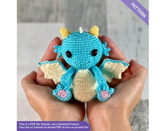 May include: A crocheted dragon in shades of blue, cream, and yellow. The dragon has large black eyes, small wings, and pink accents on its feet. The image includes the text "PATTERN" and a disclaimer about the product.