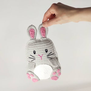 May include: A gray crocheted bunny-shaped item with pink accents. The bunny has pink inner ears, paws, and a pink nose. Black embroidered whiskers and eyes complete the design. The item is held by a hand.