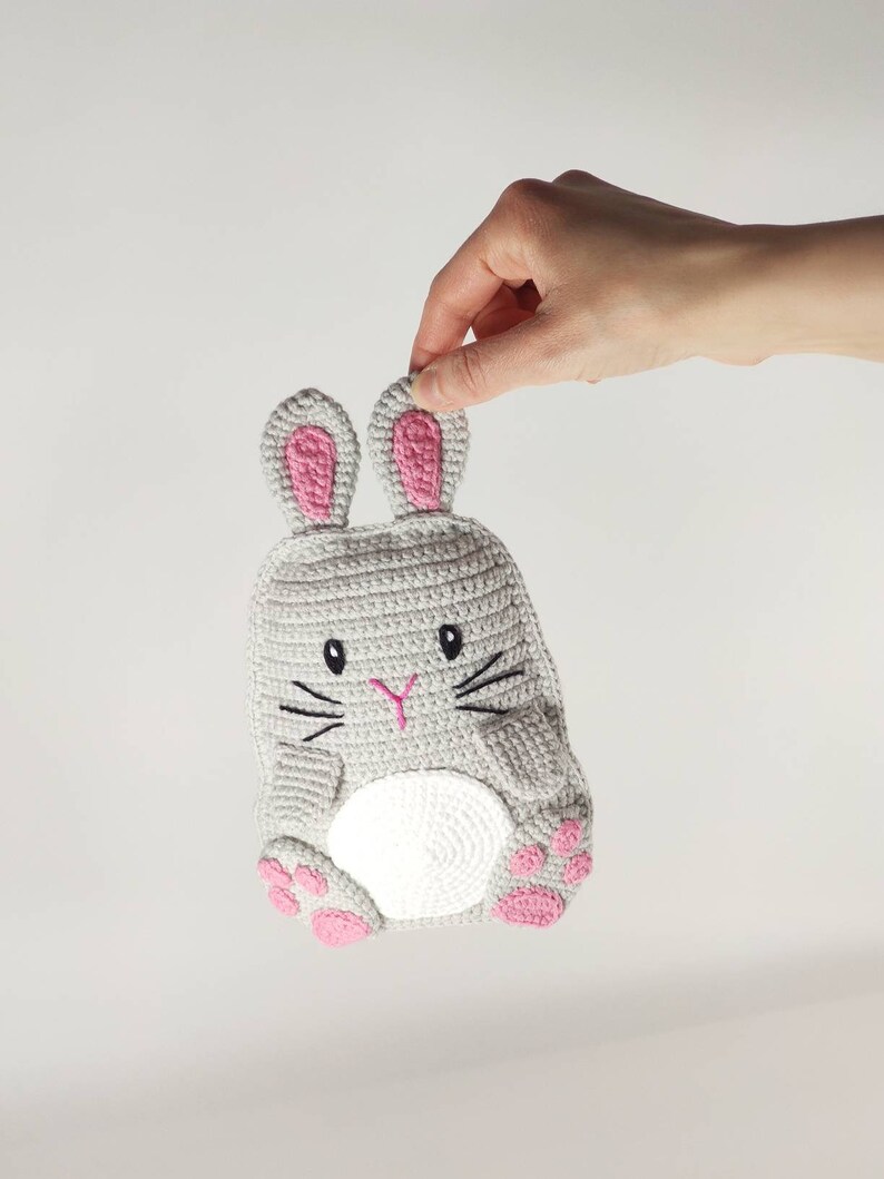May include: A gray crocheted bunny-shaped item with pink accents. The bunny has pink inner ears, paws, and a pink nose. Black embroidered whiskers and eyes complete the design. The item is held by a hand.