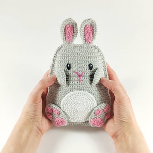 May include: A gray crochet bunny-shaped backpack with pink accents on the ears and feet. The bunny has black eyes and a pink embroidered nose. The backpack is held by two hands.