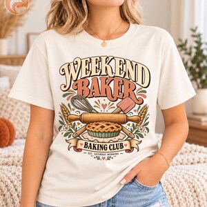 May include: A cream-colored t-shirt featuring a graphic with the text "WEEKEND BAKER" and "BAKING CLUB EST. SATURDAY MORNING." The design includes a whisk, a spatula, a rolling pin, wheat stalks, and a pie, all in muted earth tones.