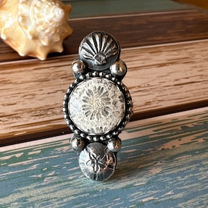 May include: A handcrafted silver ring with a unique design. The ring features a central circular stone with a white floral pattern, framed by a beaded border. Above and below are seashell and sand dollar motifs, adding a coastal theme. The ring is set against a wooden background.