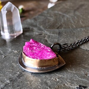 May include: A pendant necklace featuring a vibrant pink crystal set in a silver and gold-tone metal frame. The necklace has a delicate chain and a unique, textured gemstone. A clear crystal point is in the background.
