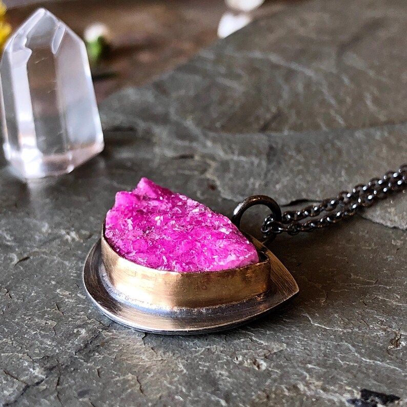 May include: A pendant necklace featuring a vibrant pink crystal set in a silver and gold-tone metal frame. The necklace has a delicate chain and a unique, textured gemstone. A clear crystal point is in the background.