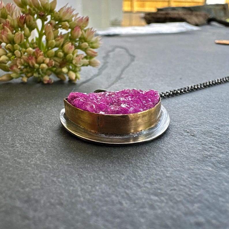 May include: A pendant necklace featuring a vibrant pink druzy stone set in a gold-toned metal bezel. The pendant rests on a silver-colored base and hangs from a delicate chain. The necklace is displayed on a dark gray surface.