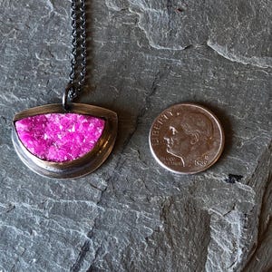 May include: A silver-toned pendant necklace with a half-moon shaped pendant filled with bright pink crystals. A U.S. dime is next to the pendant for size comparison. The necklace chain is dark gray.