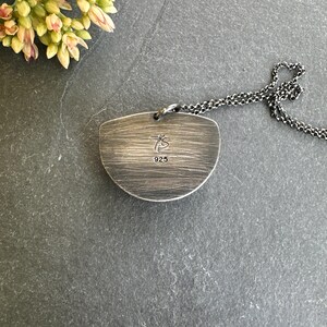 May include: A silver-toned pendant necklace with a textured, half-moon shaped pendant. The pendant has the text "TS" and "925" engraved on it. The chain is a dark, linked design. The necklace is displayed on a dark gray surface.