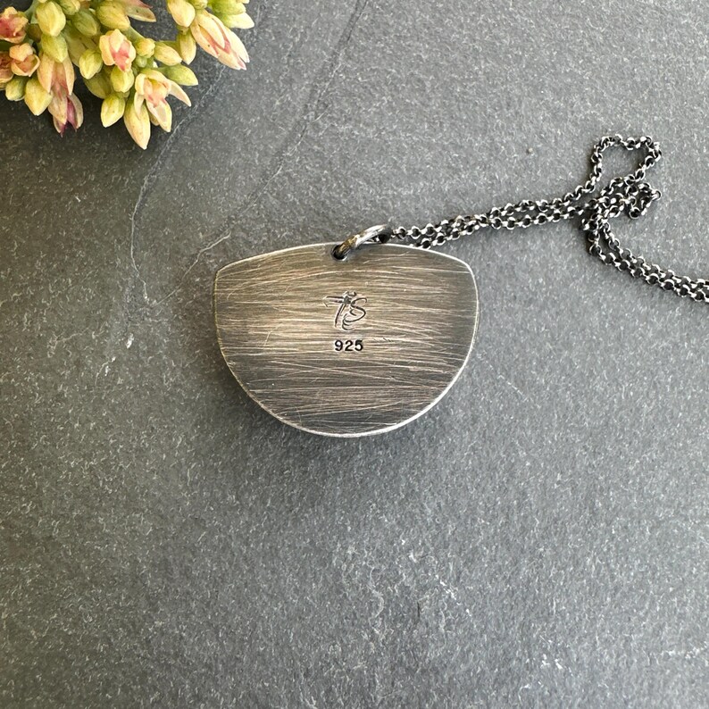 May include: A silver-toned pendant necklace with a textured, half-moon shaped pendant. The pendant has the text "TS" and "925" engraved on it. The chain is a dark, linked design. The necklace is displayed on a dark gray surface.