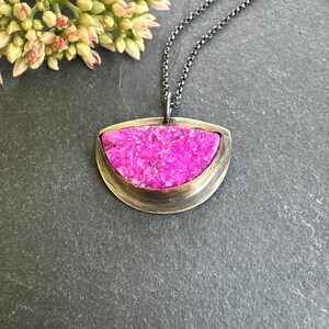 May include: A silver-toned pendant necklace featuring a vibrant pink, textured stone set in a gold-toned bezel. The pendant hangs from a delicate chain. The necklace is displayed on a dark gray surface, with a sprig of flowers in the upper left corner.