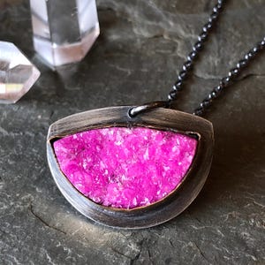 A silver-toned pendant necklace featuring a vibrant pink, irregularly shaped stone set within a gold filled bezel. The stone has a textured, granular appearance. The pendant hangs from a dark, oxidized chain. Handmade oxidized silver necklace and pendant, lightly hand polished contrast the bright pink druzy stone.  Made with the utmost care and attention to detail. Thoughtfully packaged and ready for gift giving.
