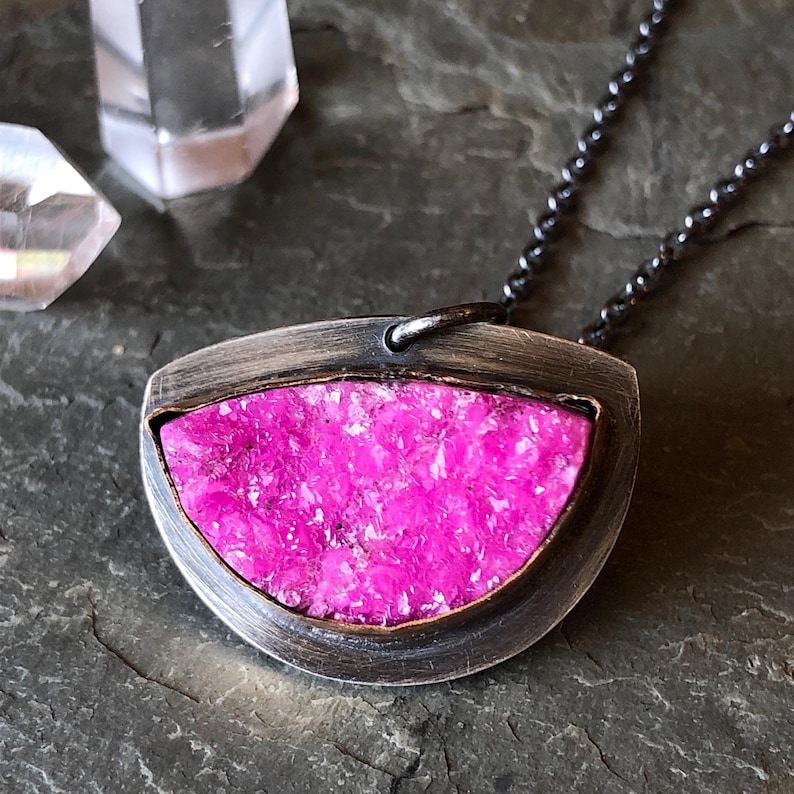 A silver-toned pendant necklace featuring a vibrant pink, irregularly shaped stone set within a gold filled bezel. The stone has a textured, granular appearance. The pendant hangs from a dark, oxidized chain. Handmade oxidized silver necklace and pendant, lightly hand polished contrast the bright pink druzy stone.  Made with the utmost care and attention to detail. Thoughtfully packaged and ready for gift giving.